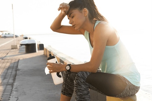 A woman sitting outdoors in athletic wear with her hand on her forehead.
