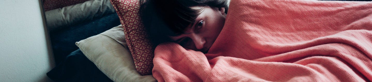 Person lying in bed under a pink blanket appearing unwell