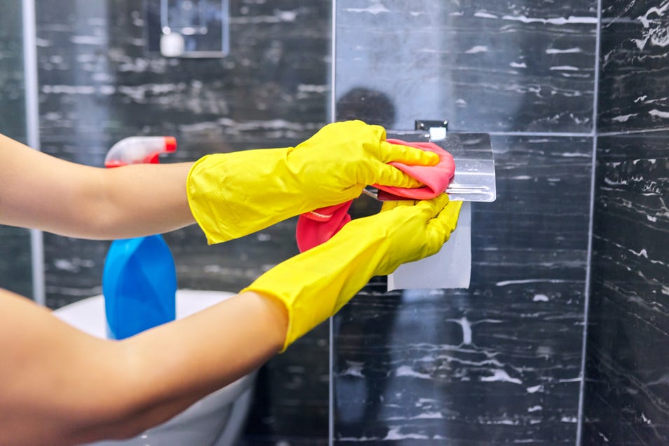 A person wearing yellow rubber gloves using a cloth to clean a toilet paper holder