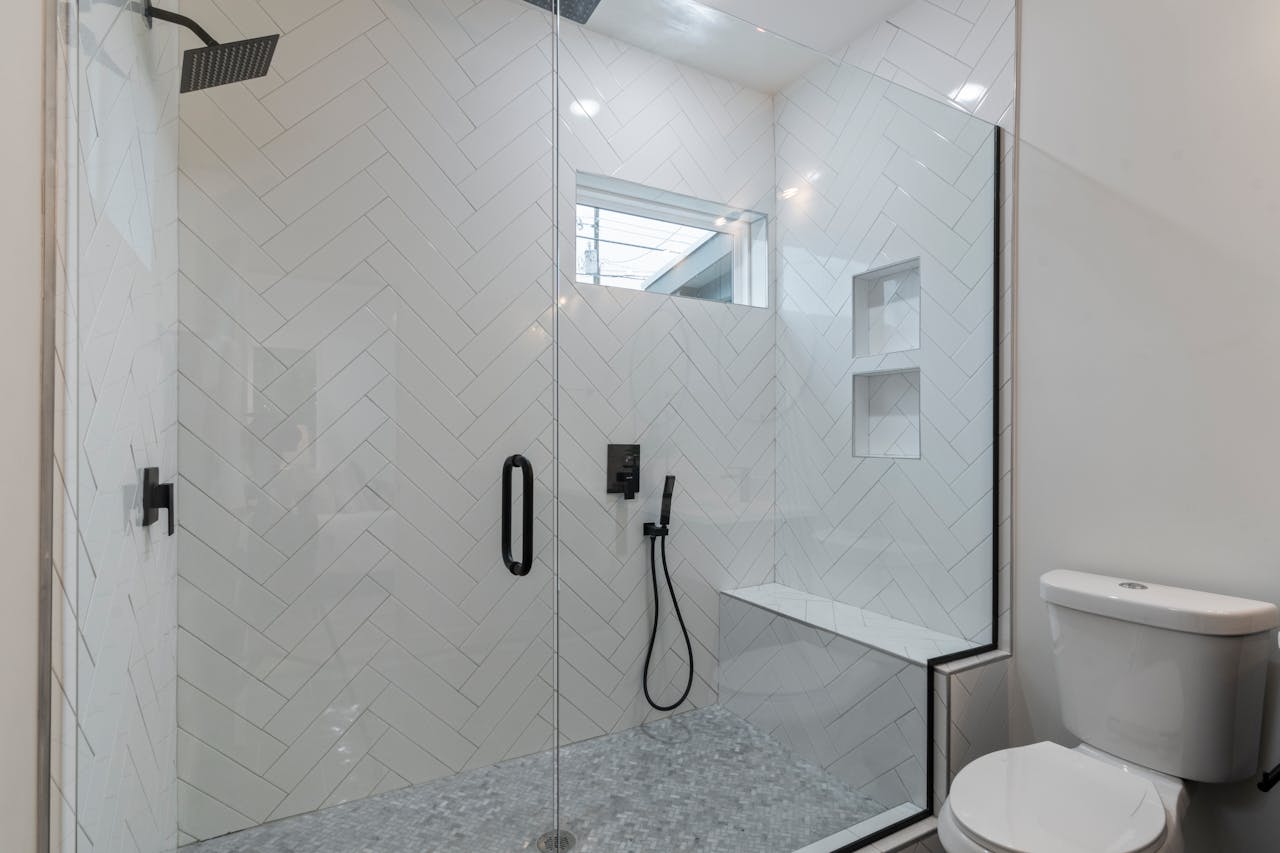 Banheiro moderno com box de vidro, azulejos brancos em padrão chevron, chuveiro preto e área de assento, ao lado de um vaso sanitário branco.