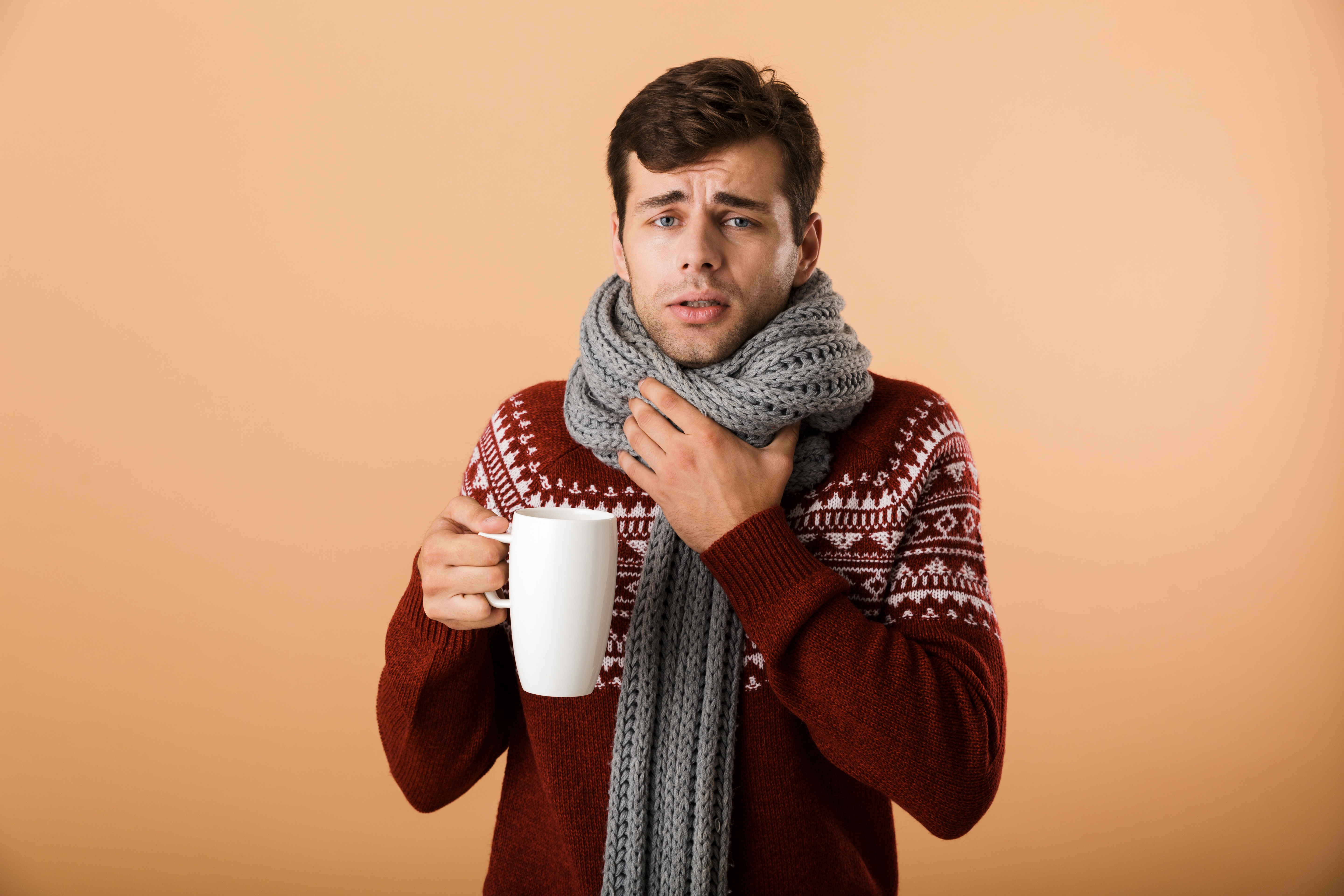 Man holding his throat, wearing a scarf, and holding a mug.
