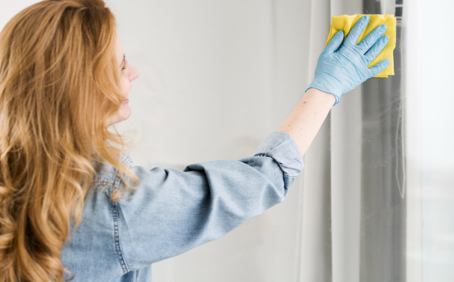 Mulher limpando espelho com pano amarelo, usando luvas azuis e camisa de jeans. 