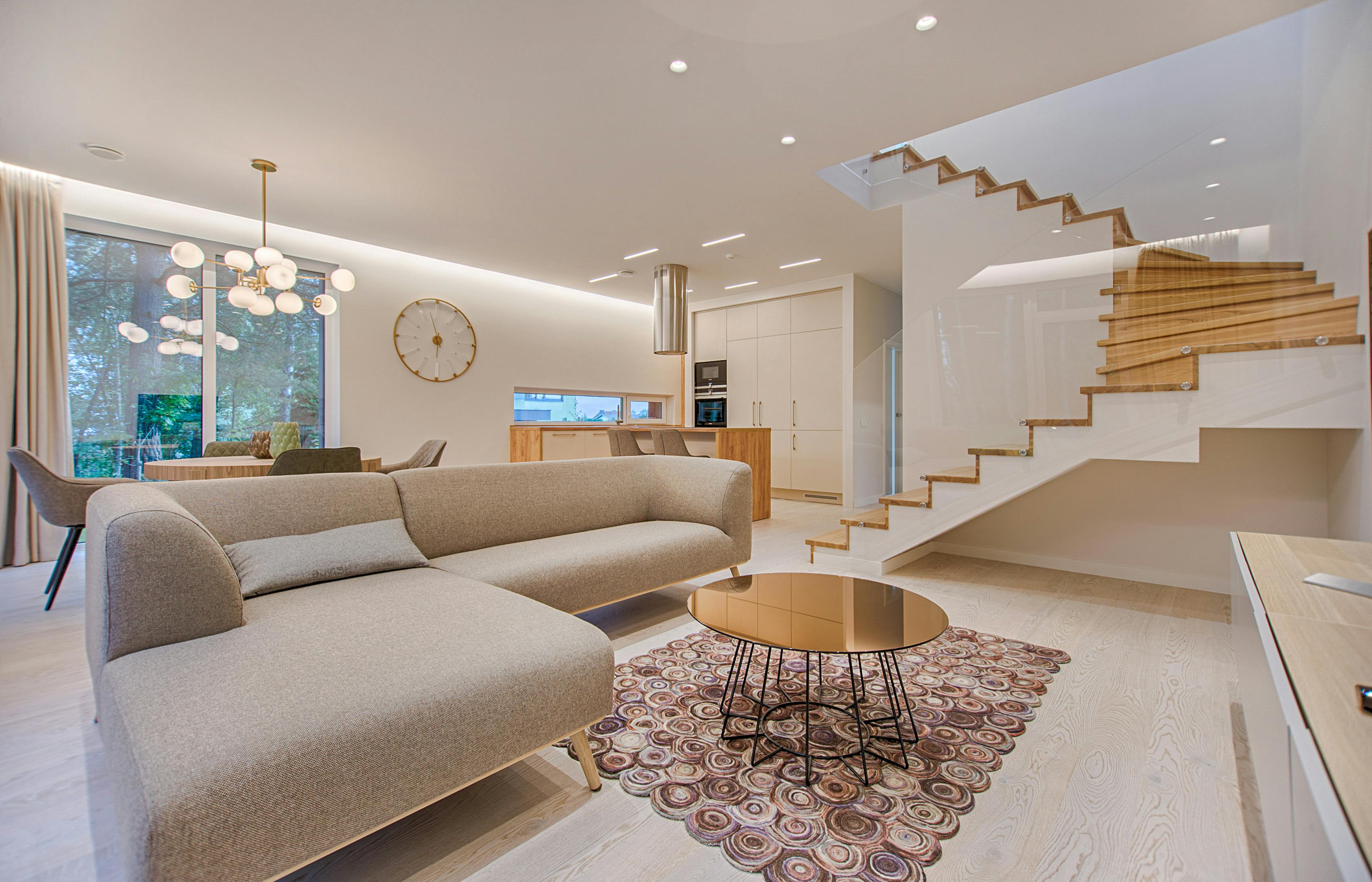 Sala de estar moderna integrada com cozinha, sofá bege, escada flutuante de madeira, mesa de centro redonda e iluminação sofisticada.