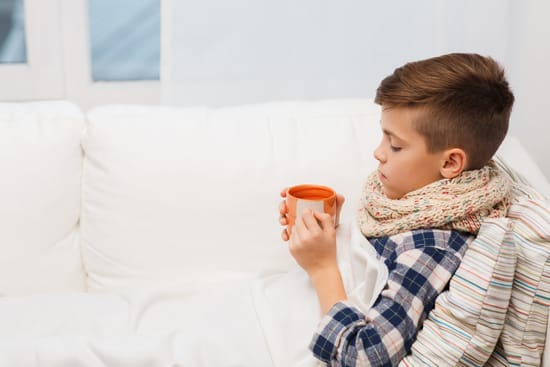 A sick boy resting under a blanket while holding a warm mug.