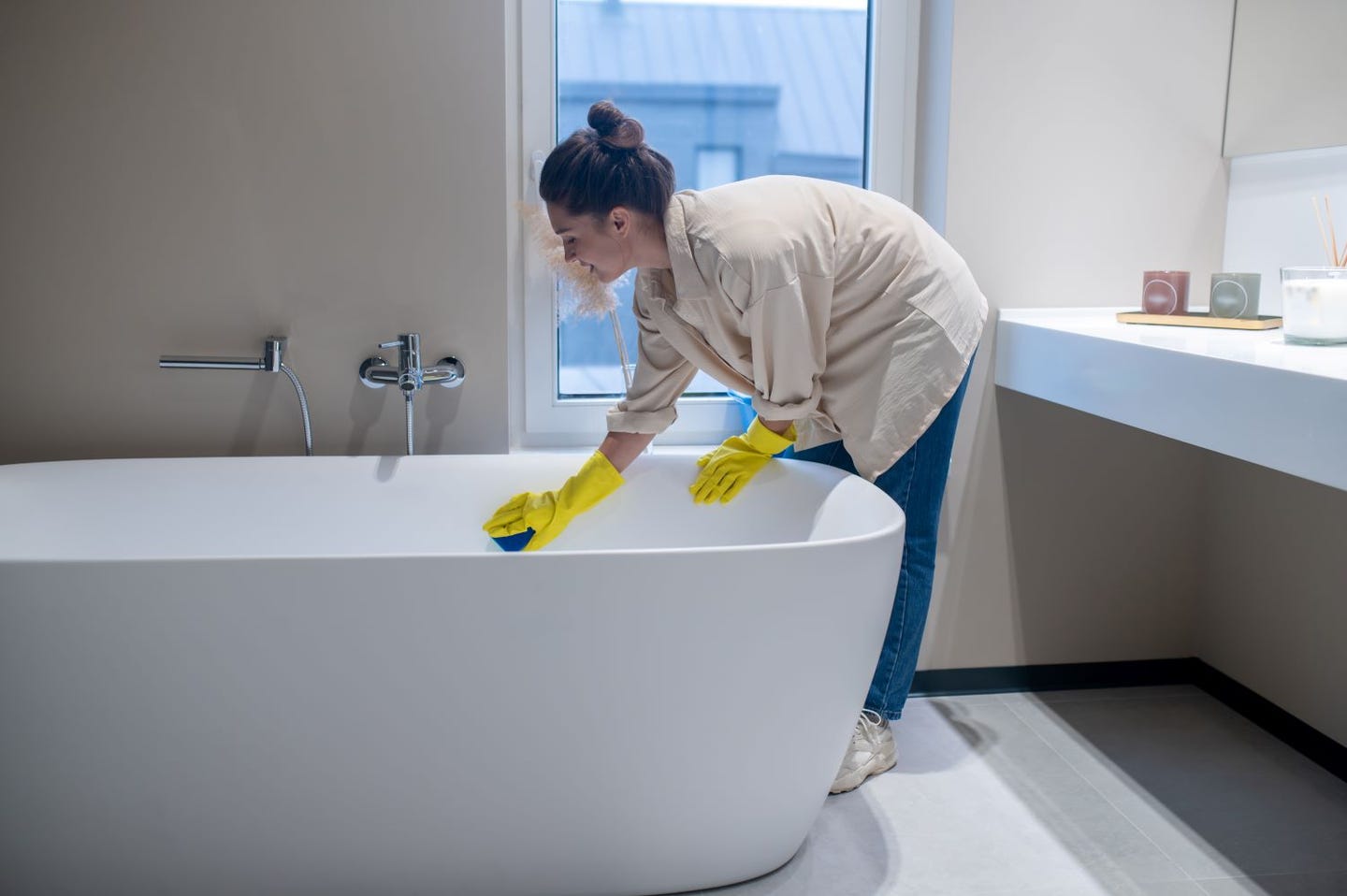 A woman wearing gloves and cleaning a bathtub with a sponge while standing