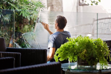 Homem limpando vidro com rodo, enquanto plantas decoram o fundo.