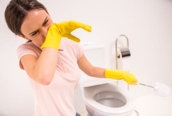 A woman wearing rubber gloves, covering her nose while cleaning a toilet with a toilet brush.