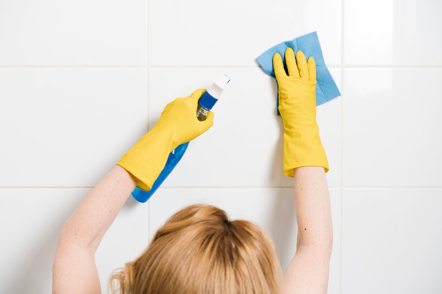Woman wearing rubber gloves holding spray bottle and cloth to clean shower tiles.
