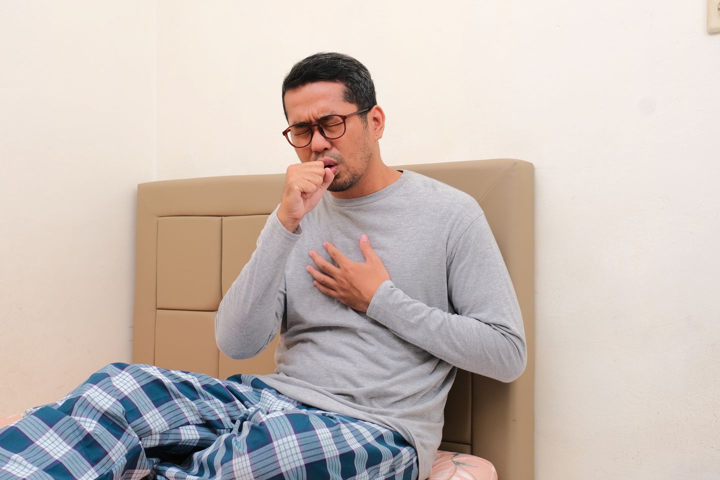 A man sitting on the his bed, covering his mouth as he coughs