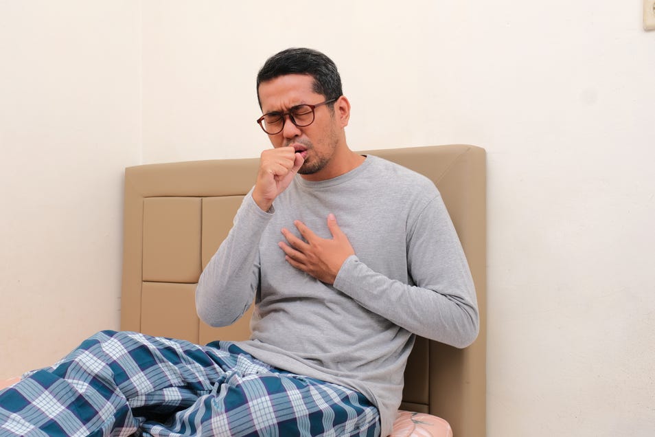 A man sitting on the his bed, covering his mouth as he coughs