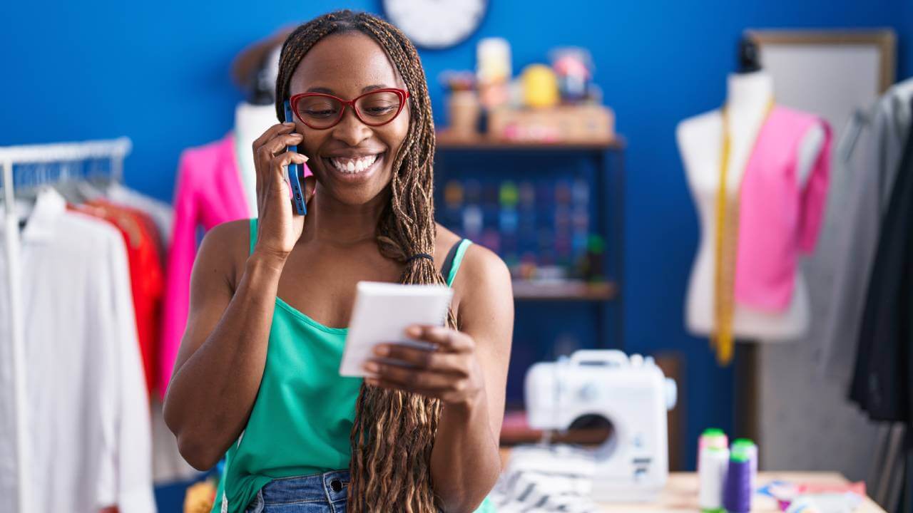 African seamstress talking on a smartphone, smiling and looking at a notebook.