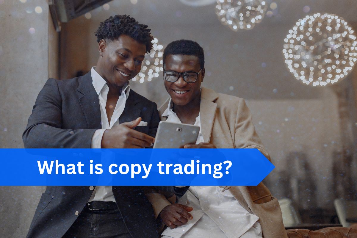 Two men in formal attire smiling while looking at a tablet in a warmly lit setting with decorative lights in the background. A blue banner with white text reads 'What is copy trading?