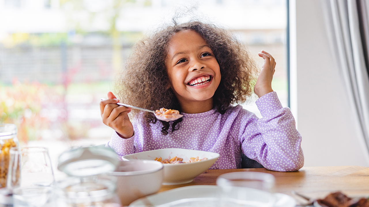 Adorable Black Girl With Curly Hair Smiling While Eating Breakfast In A Bright Kitchen