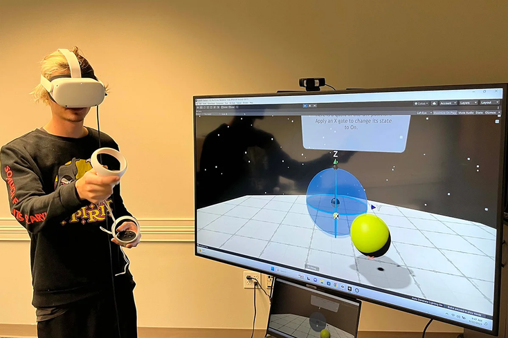 A student wearing a VR headset and holding a controller stands next to a screem showing an illustration of a quantum gate A student wearing a VR headset and holding a controller stands next to a screem showing an illustration of a quantum gate