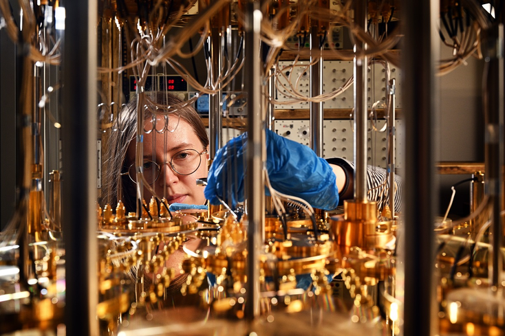A woman in a lab coat wearing blue gloves and glasses works on a golden quantum computer 