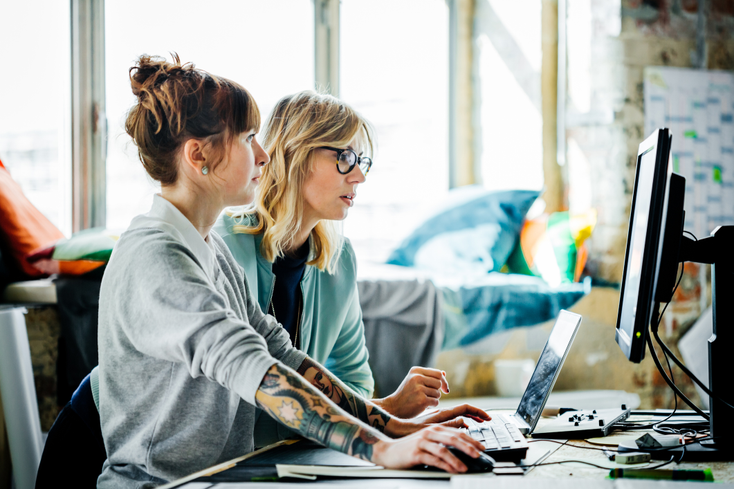 Two women collaborating at a computer. One has tattoos Two women collaborating at a computer. One has tattoos