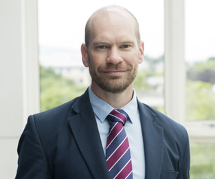 A portrait image of Andrew Mitchell with short hair and bears, wearing a suit