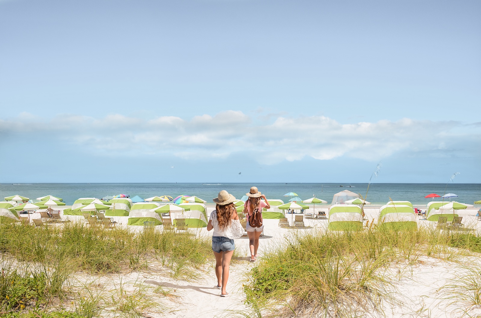 Two women walk on Clearwater beach on a summer holiday. Beach chairs and umbrellas are on the white sand and the ocean is in the background.