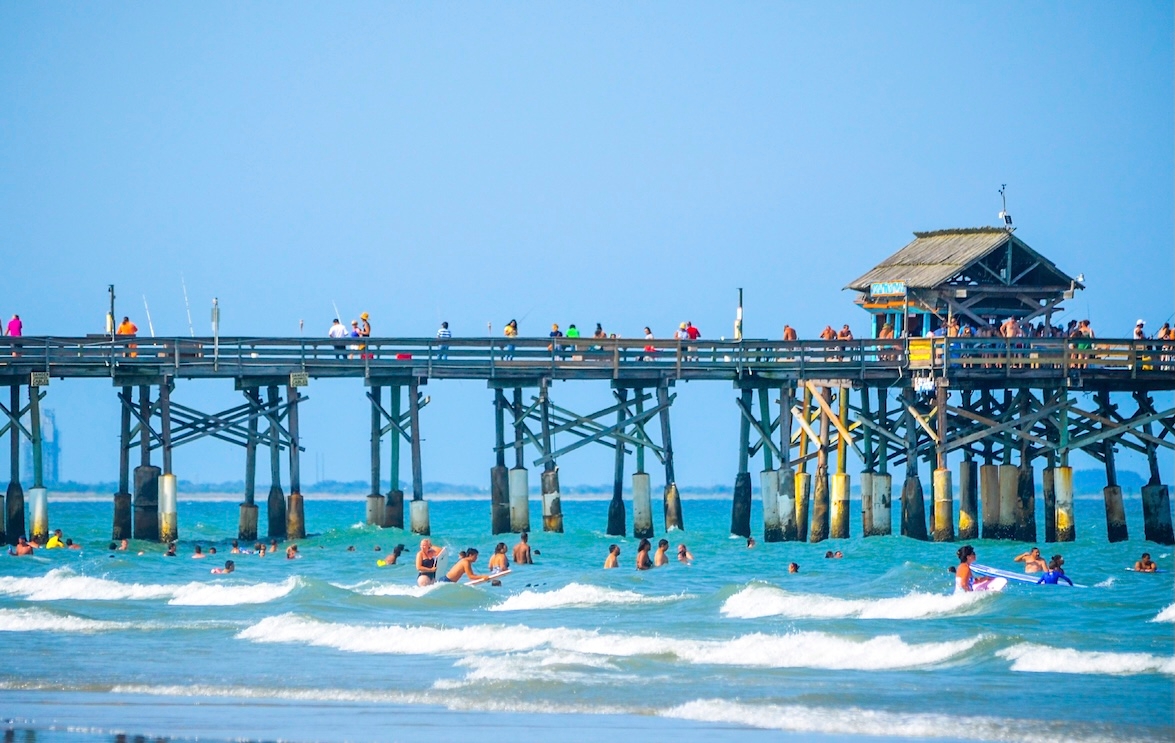 A pier at Cocoa Beach with people swimming in the ocean under a clear blue sky.