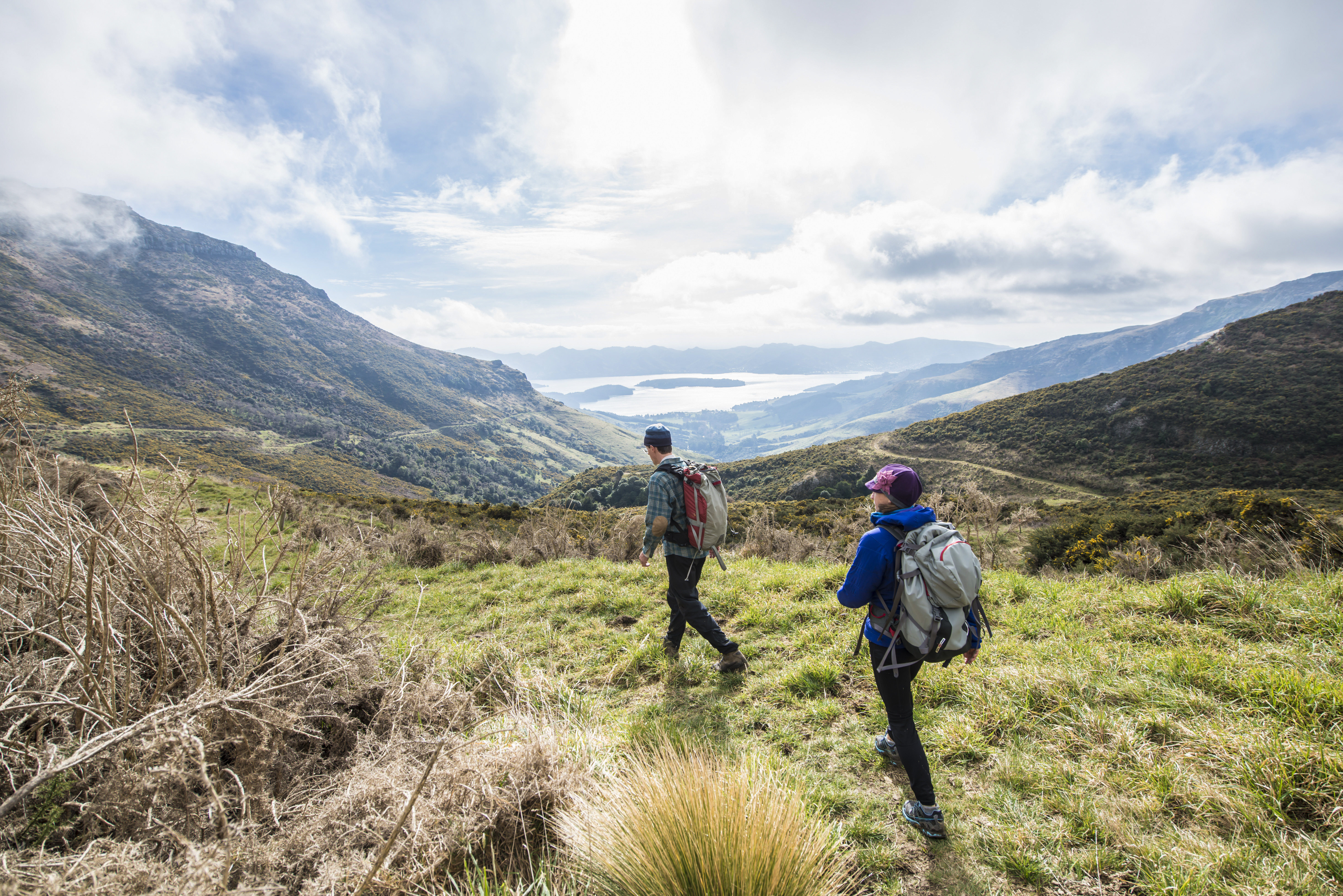 Un hombre y una mujer con mochilas caminan por un sendero cubierto de hierba en la ladera, hacia un valle pintoresco en Nueva Zelanda, con un gran lago y montañas a lo lejos bajo un cielo nublado.