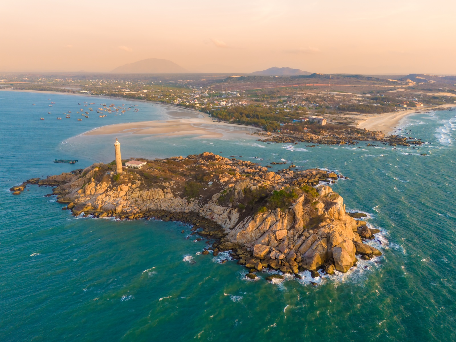Aerial view of a lighthouse at sunset over the ocean, located in Mũi Né, Vietnam, with vibrant colours reflecting on the water.