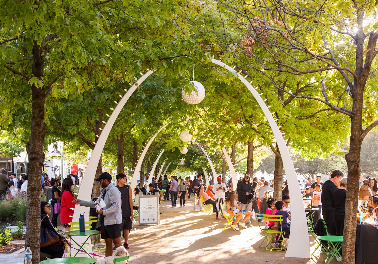 A crowd of people seated at tables under trees in the Dallas Arts District enjoy the outdoor atmosphere