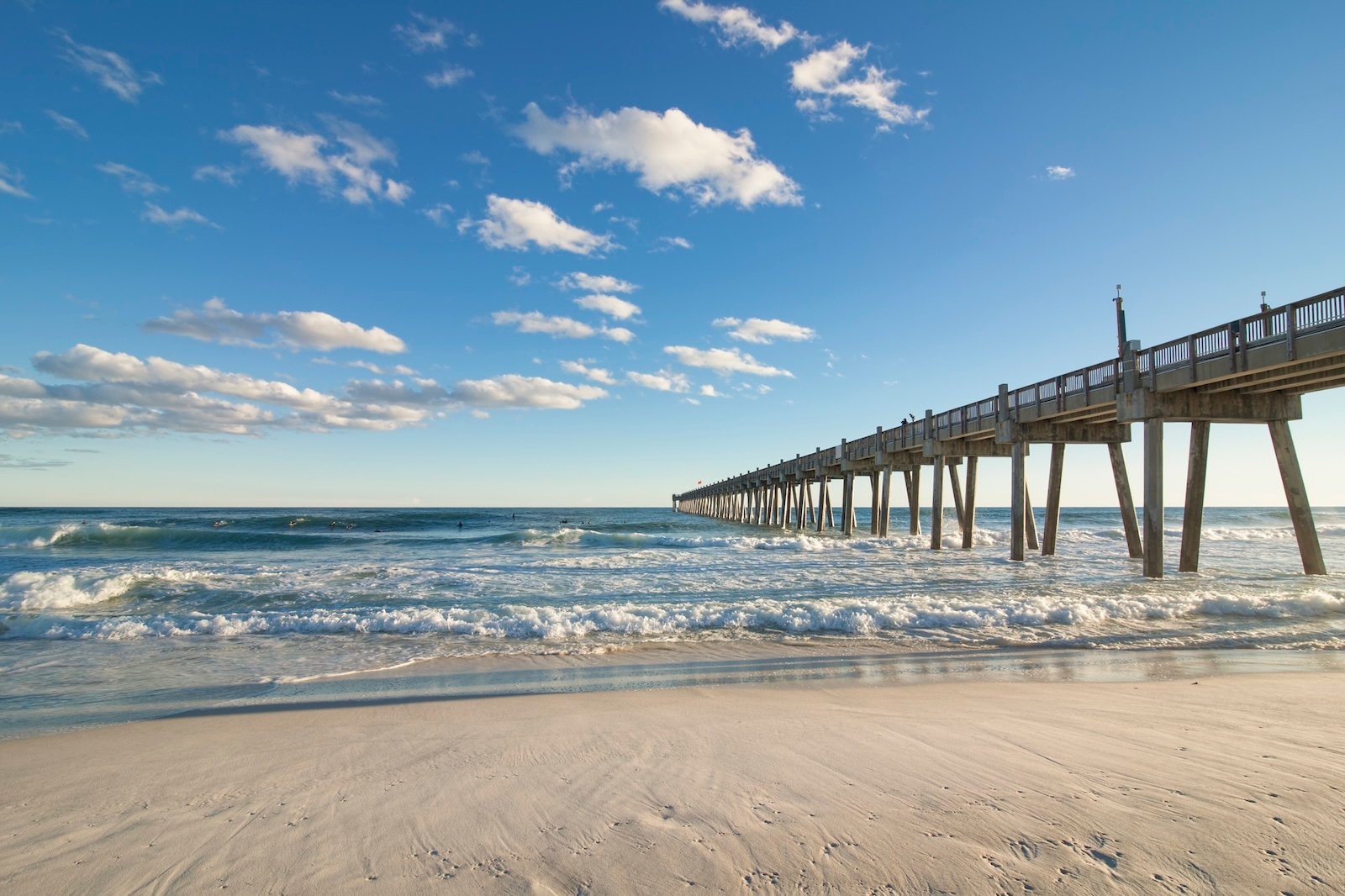  A pier at Pensacola Beach with waves crashing against it under a cloudy, yet bright blue sky