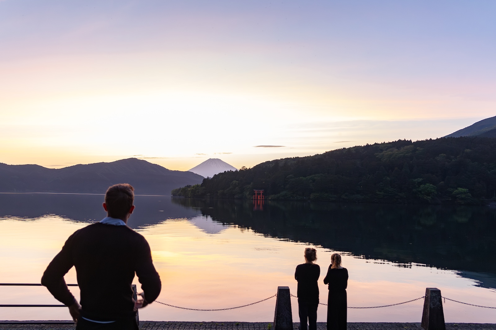 Some silhouette figures stand on a dock, gazing at the sunset over Lake Hakone, creating a serene and romantic atmosphere.