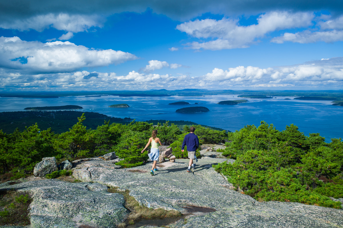 two hikers stroll along a rocky hill in Acadia National Park, Maine, surrounded by stunning natural scenery.
