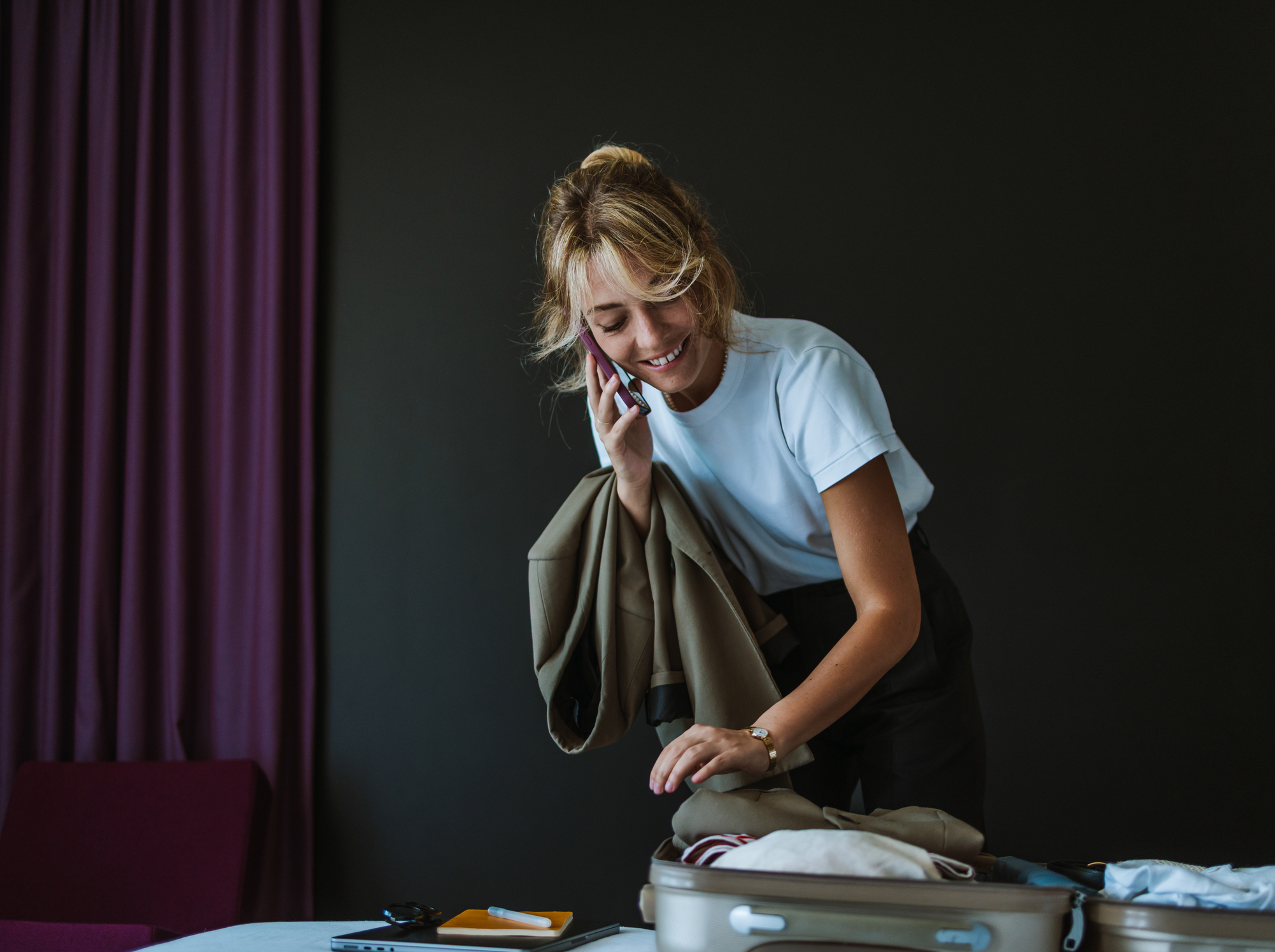 A woman smiling and holding a mobile phone while folding clothes into an open suitcase on a table