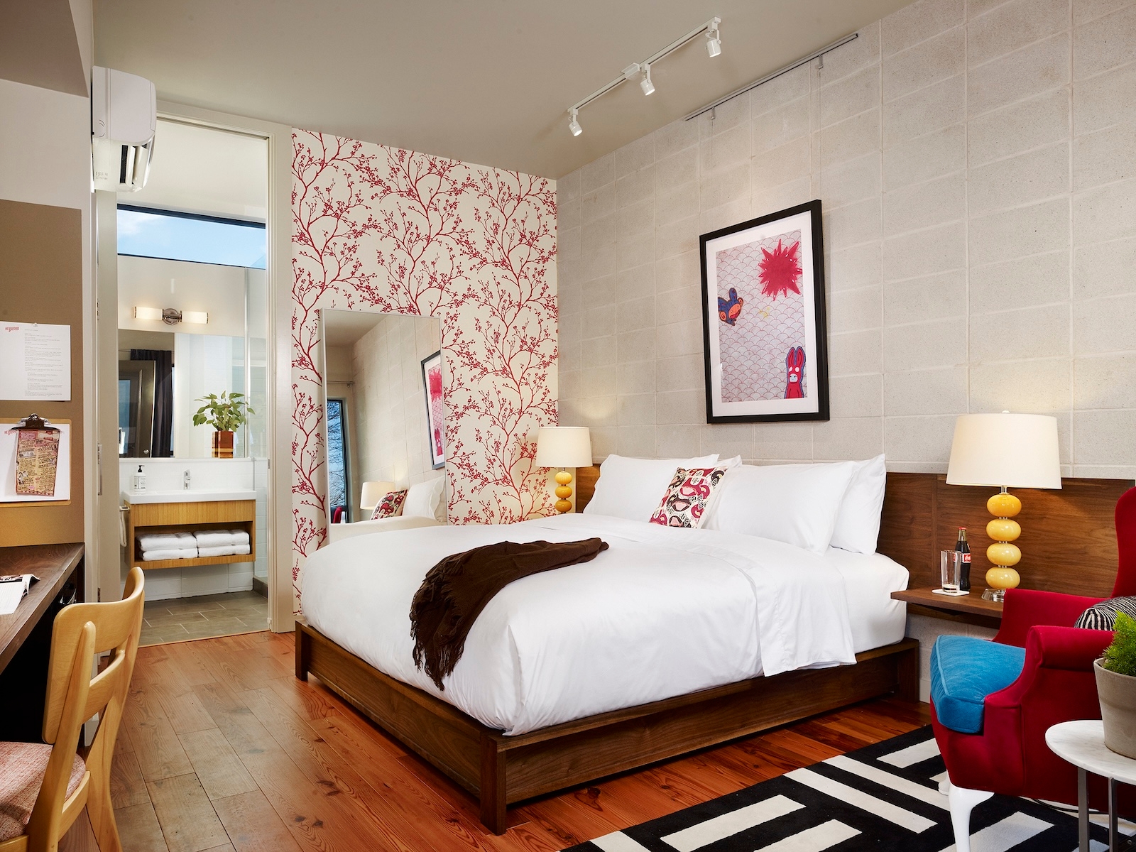A hotel room at red-accented Heywood Hotel featuring a bed, desk, chair, and a floral-patterned wall.