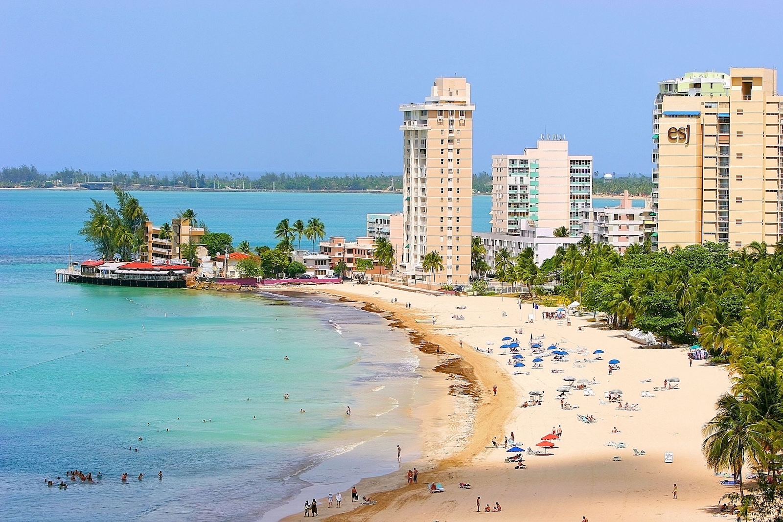 A beach at Parque Isla Verde, featuring numerous people enjoying the sun and buildings lining the shore