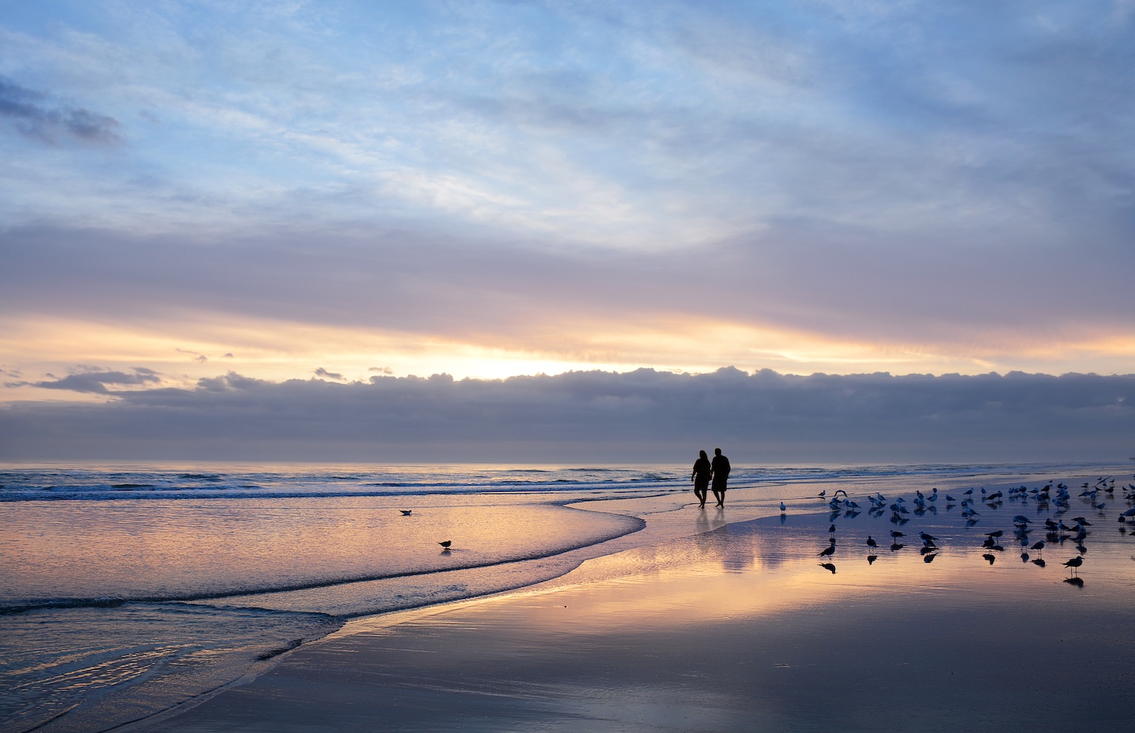 A couple strolls hand in hand along Daytona Beach at sunset, with vibrant colours reflecting on the water.