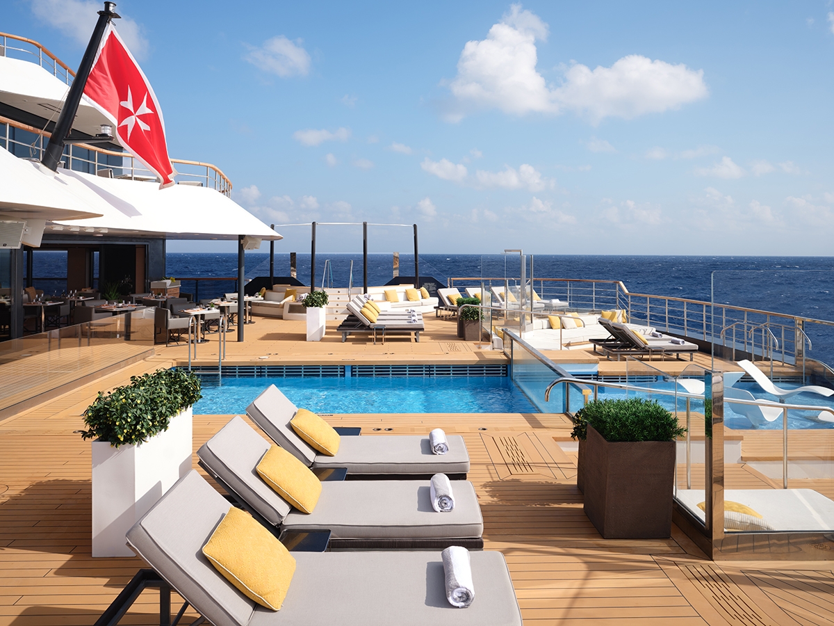 A large pool on a luxurious yacht from the Ritz Carlton Yacht Collection. There are grey sun beds with yellow cushions in the foreground.