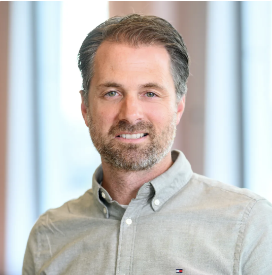 A close-up portrait of a smiling adult man with short dark hair and light facial hair, wearing a light gray button-down shirt. The background is softly blurred, suggesting an indoor setting with window light.