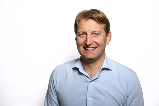 Studio-style head-and-shoulders portrait of an adult man smiling at the camera, wearing a light blue button-down shirt against a plain white background.