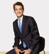 Man smiling with dark hair in a dark suit with a white shirt and light blue tie sitting on a stool with hands place in front of him