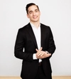 Man smiling with dark hair in a dark suit with a white shirt standing with hands crossed