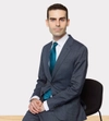 Man smiling with dark hair in a dark suit sitting on a stool with both hands placed in front