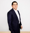 Man smiling with glasses and dark hair in a dark suit with a white shirt standing with one hand in his pocket