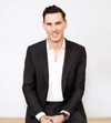 Man smiling with dark hair in a dark suit sitting on a stool with both hands placed in front