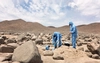 Three workers in protective gear work in a field of boulders.