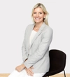 Woman smiling with light hair in a light suit with a white shirt sitting on a stool with hands wresting in front of her