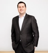 Man smiling with dark hair in a dark suit with a white shirt standing with both hands in pockets