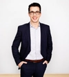 Man smiling with dark hair  and glasses in a dark suit with a white shirt standing with both hands in his pocket