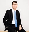 Man smiling with dark hair in a dark suit sitting on a stool with one hand in pocket