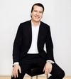 Man smiling with dark hair in a suit with a white shirt sitting on a stool with hands wresting in front
