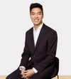 Man smiling with dark hair in a dark suit sitting on a stool with both hands placed in front