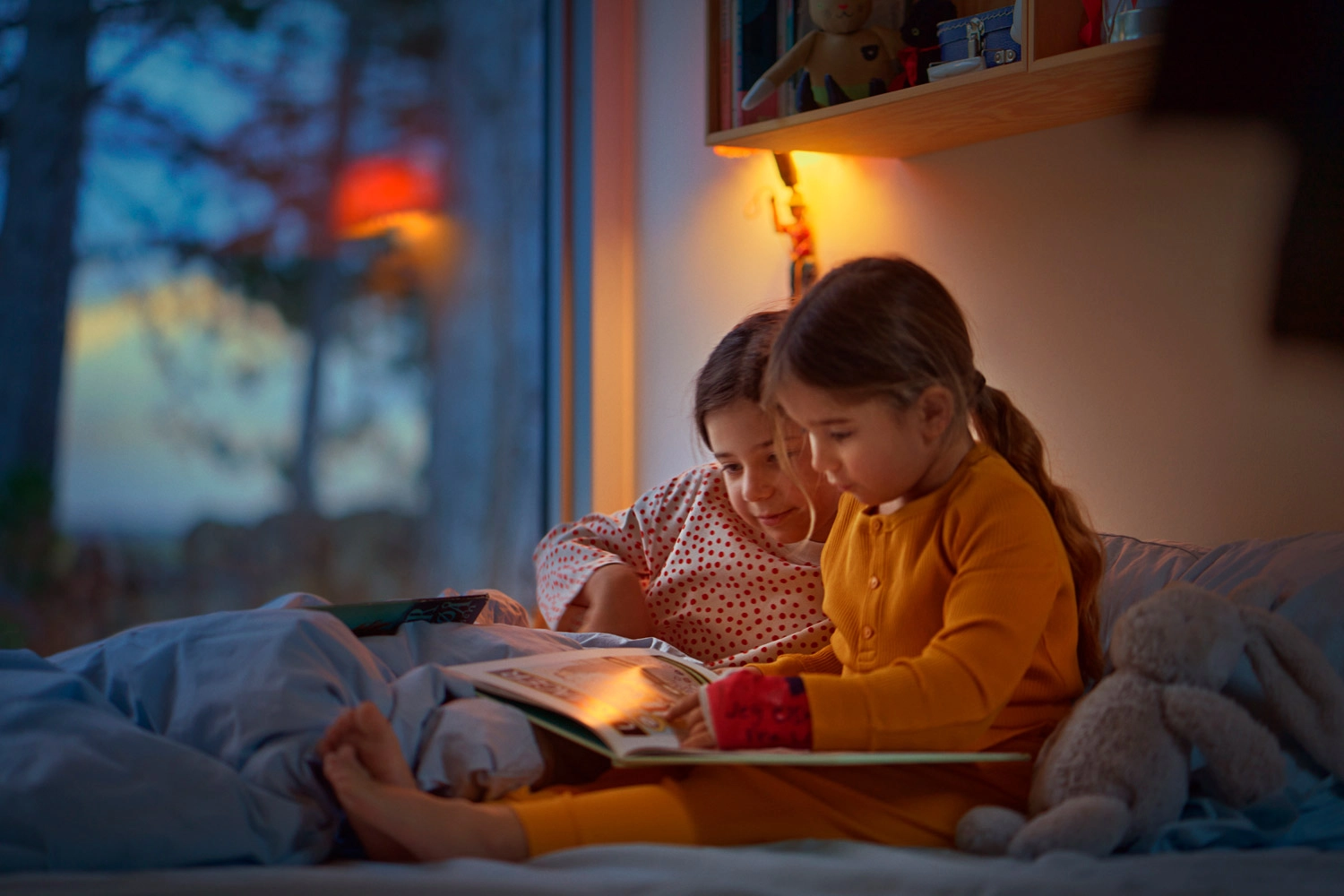 To børn sidder i en seng om aftenen og kigger i en bog med en læselampe hængende over dem
