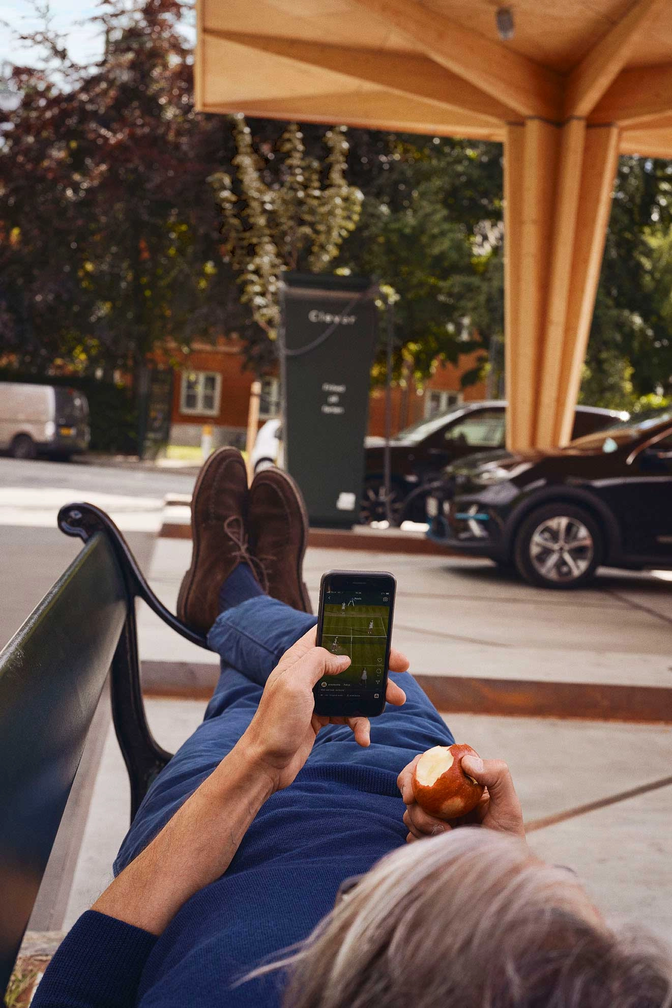 En mand ligger på en bænk og kigger på sin telefon imens hans bil holder til opladning